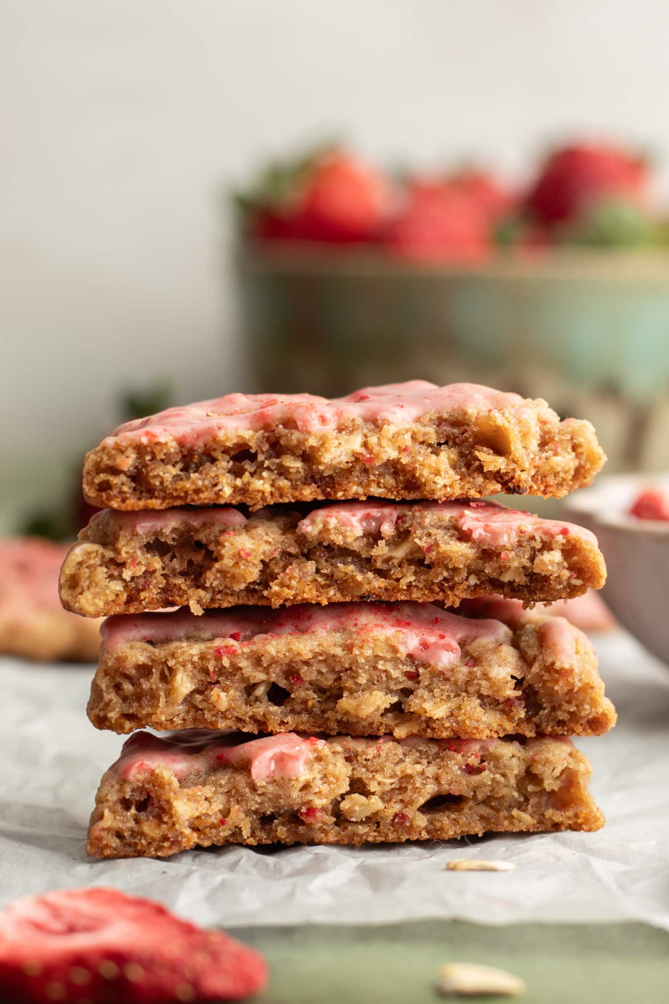 A close-up stack of halved strawberry oatmeal cookies showing their soft, chewy centers and visible oats, with strawberry glaze on top. A bowl of fresh strawberries is softly blurred in the background.