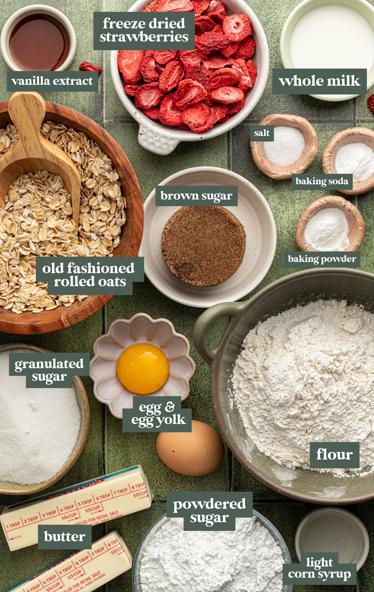 Flat lay image of labeled ingredients for strawberry iced oatmeal cookies, including old fashioned rolled oats, freeze dried strawberries, vanilla extract, whole milk, brown sugar, granulated sugar, egg and egg yolk, flour, butter, powdered sugar, baking soda, baking powder, salt, and light corn syrup. Each ingredient is neatly arranged in bowls or dishes on a green tile background.