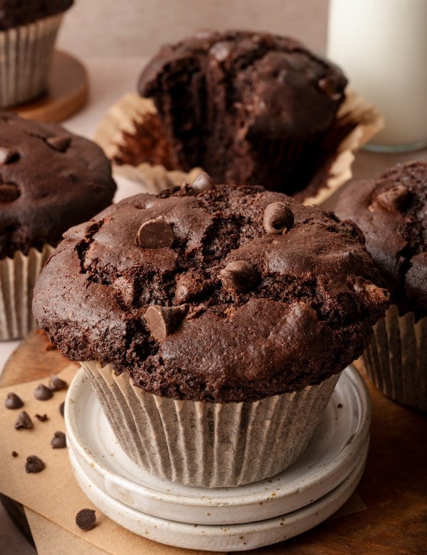 Image of double chocolate bakery-style muffin in paper liner on top of two mini serving plates and 4 muffins in the background.