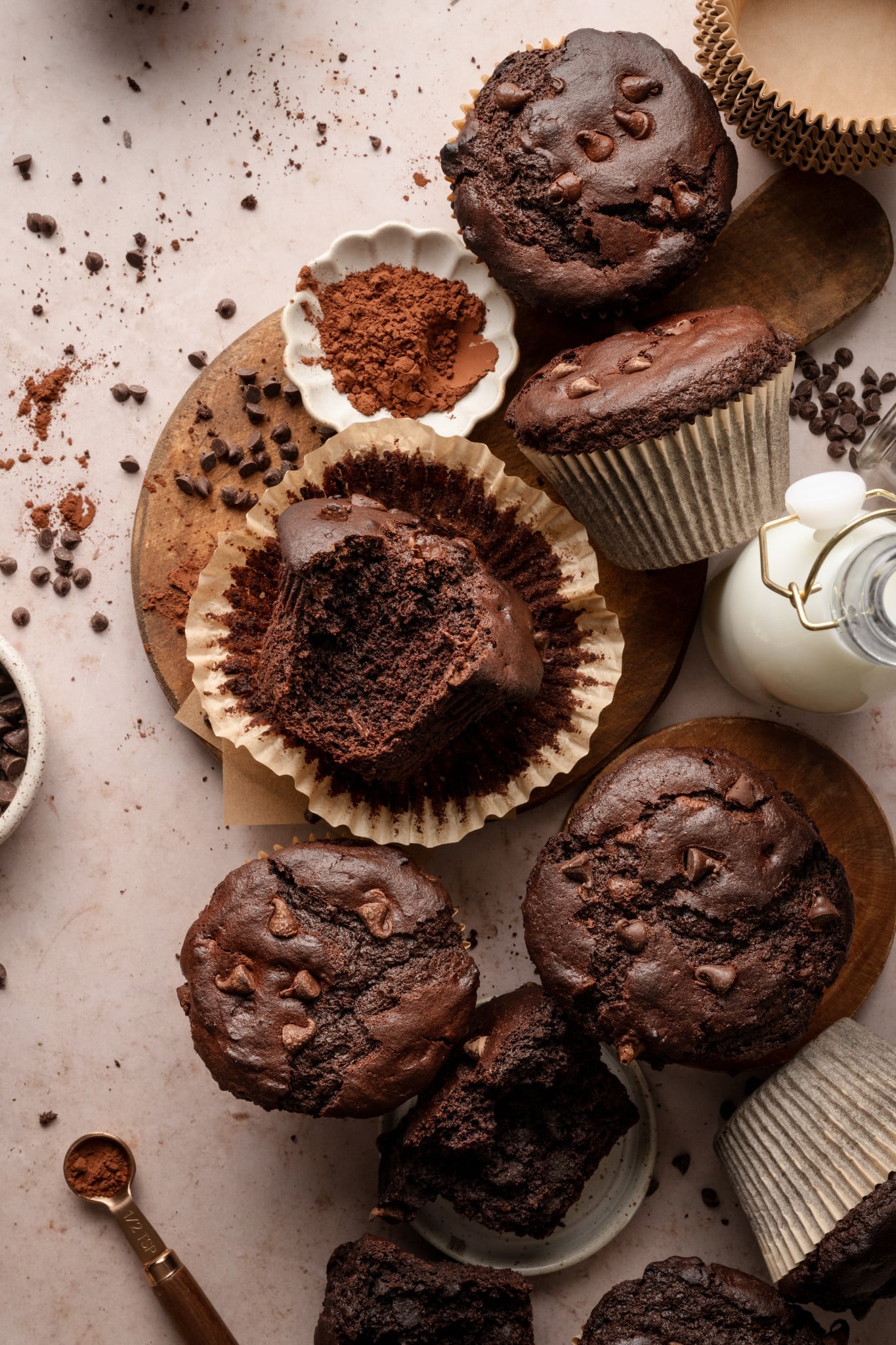 Copycat costco double chocolate muffins on top of counter top with buttermilk and cocoa powder on board. One of the muffins is unwrapped from paper liner and bite taken out of the center.