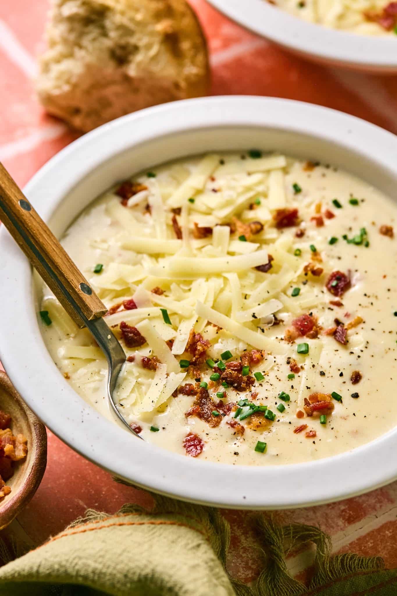 A comforting bowl of homemade potato soup, with visible chunks of potato and a creamy texture, garnished with cheese and bacon, with a torn piece of bread on the side.
