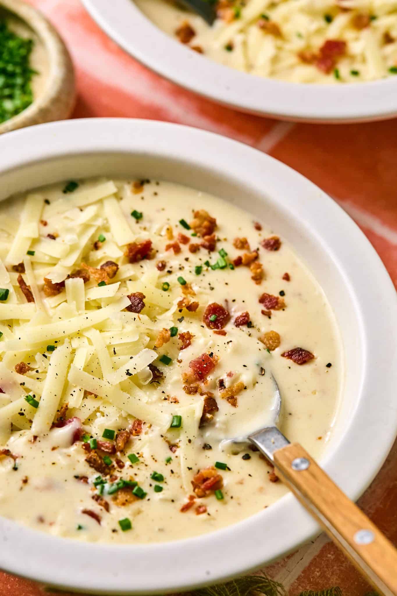 A creamy bowl of baked potato soup topped with crispy bacon, shredded cheese, and fresh chives, served in a white bowl with a wooden-handled spoon.