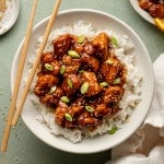Plate of panda express orange chicken in bowl on top of white rice garnished with green onions sesame seeds with pair of chopsticks on the side.