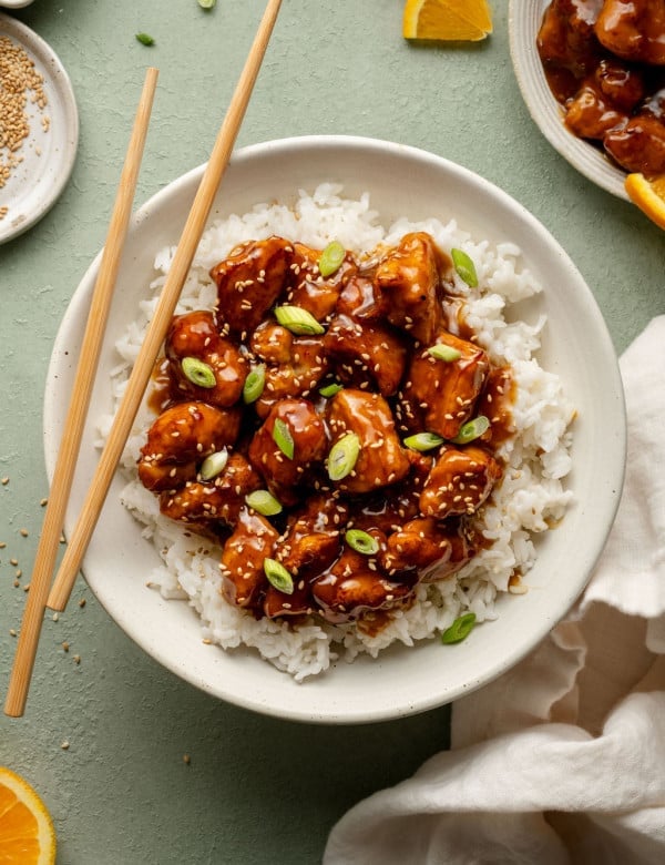 Plate of panda express orange chicken in bowl on top of white rice garnished with green onions sesame seeds with pair of chopsticks on the side.