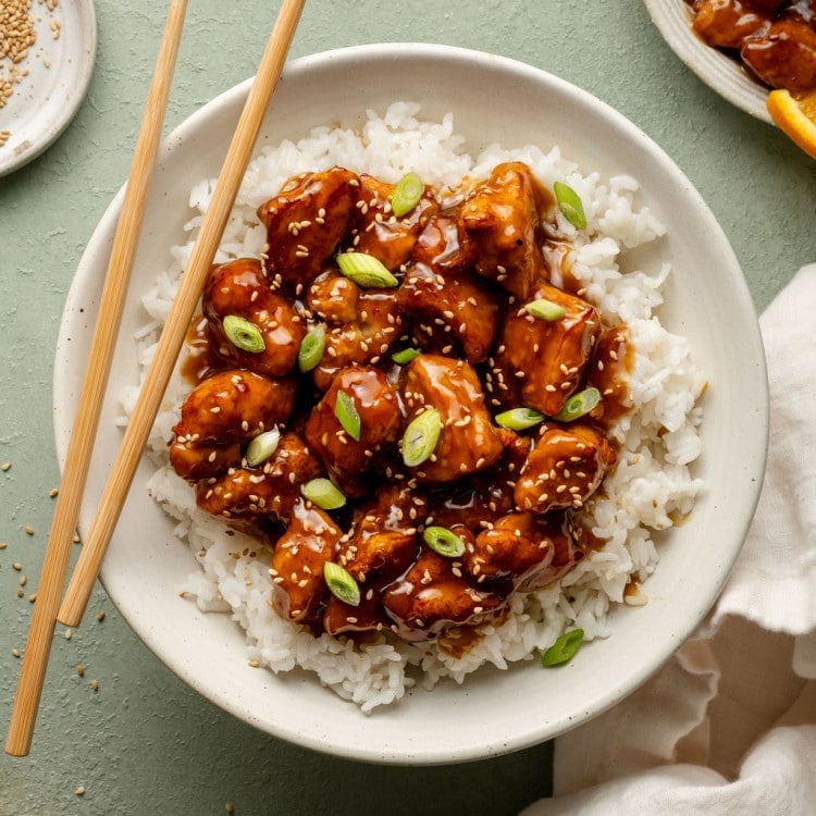 Plate of panda express orange chicken in bowl on top of white rice garnished with green onions sesame seeds with pair of chopsticks on the side.