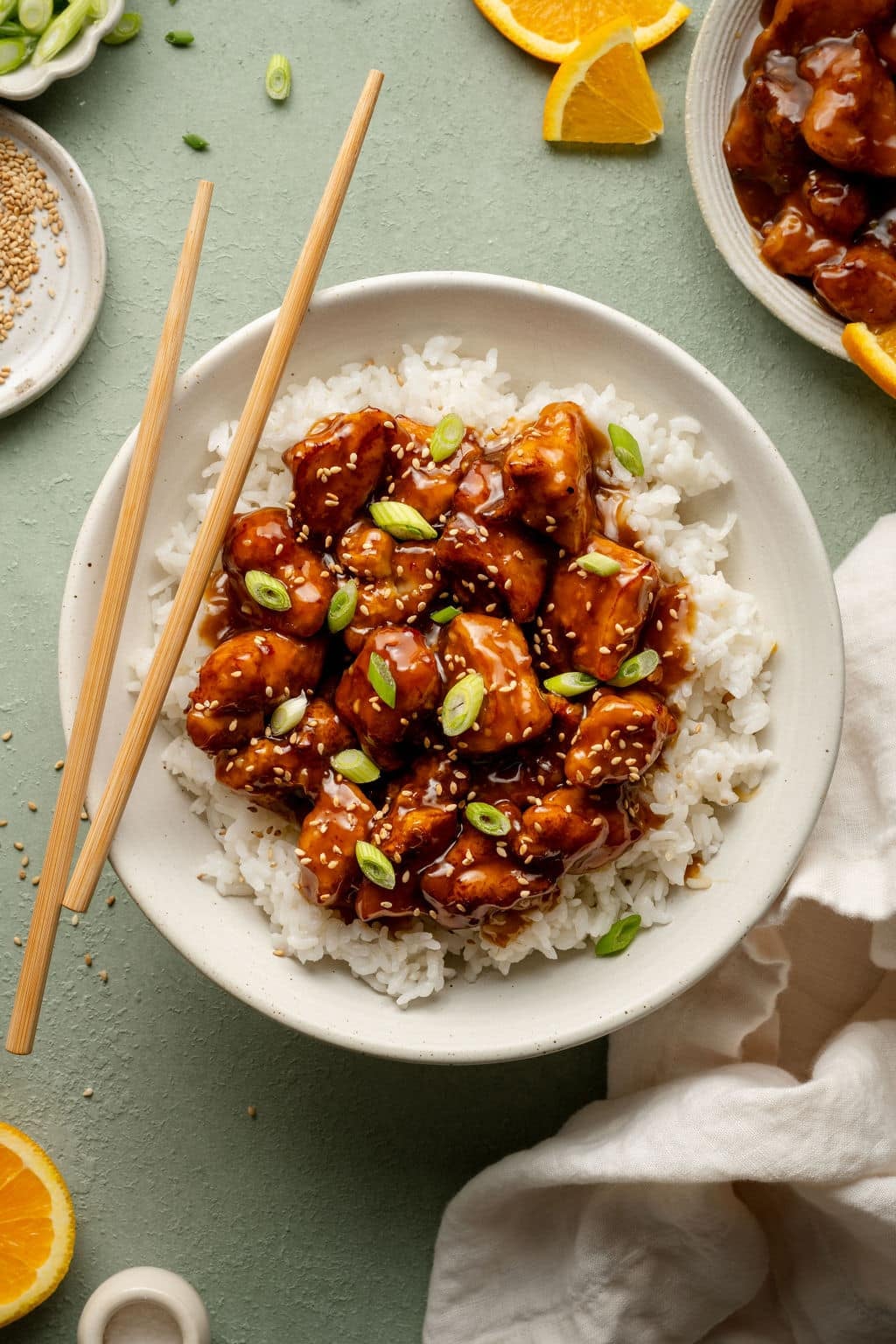 Plate of panda express orange chicken in bowl on top of white rice garnished with green onions sesame seeds with pair of chopsticks on the side.