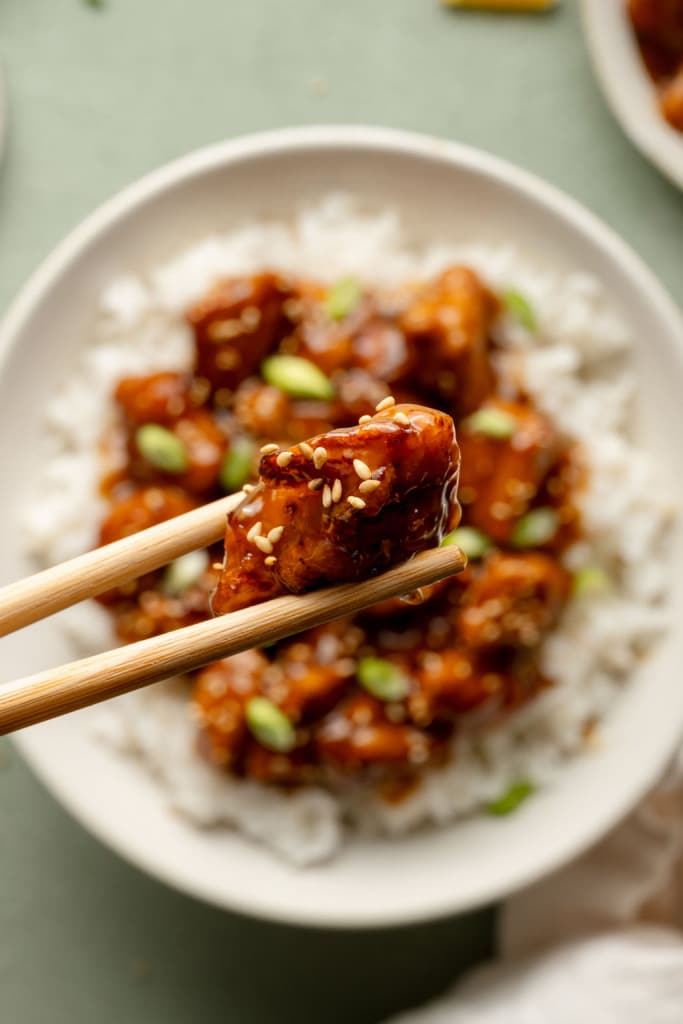 Upclose shot of chopsticks holding a piece of orange chicken. A plate of the chicken and rice is below the chopsticks on green background.