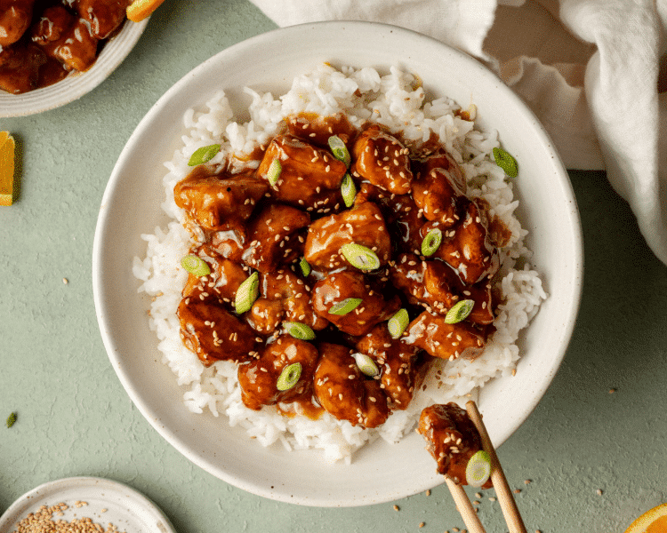 Final shot of finished orange chicken on top of white rice in a bowl. Chicken is garnished with sesame seeds and green onions and a pair of chopsticks is holding on piece of chicken.