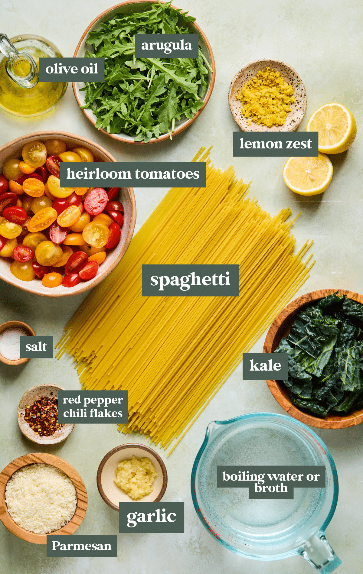 Flat lay of ingredients for Meghan Markle's one-skillet spaghetti, including uncooked spaghetti, heirloom cherry tomatoes, kale, arugula, garlic, olive oil, lemon zest, grated Parmesan, red pepper chili flakes, salt, and a measuring cup of boiling water or broth, all arranged neatly on a light surface.




