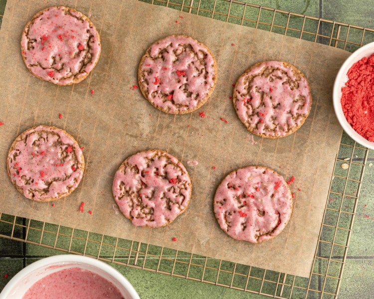 Strawberry-glazed oatmeal cookies resting on a parchment-lined rack, some sprinkled with extra strawberry powder for garnish.