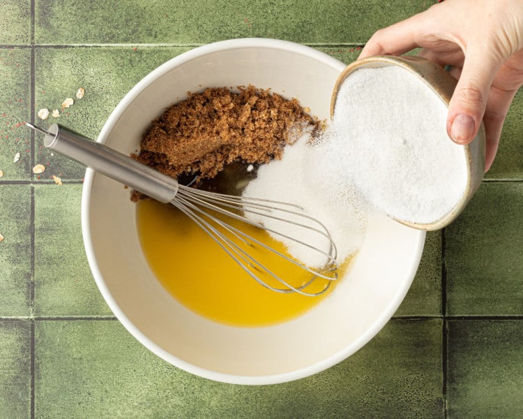 Adding granulated sugar to a bowl with brown sugar and melted butter, with a whisk resting in the mixture.