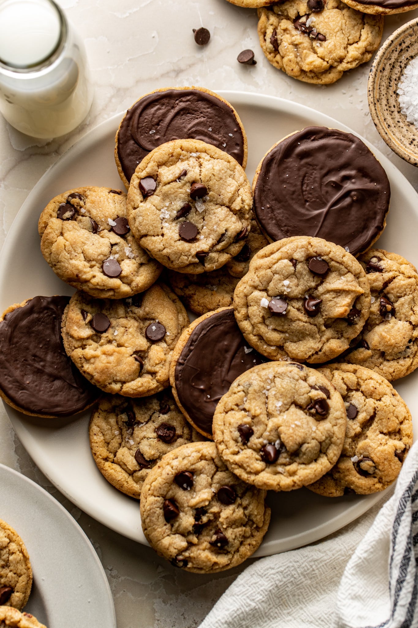 Plate of chocolate dipped cookies with some of the cookies turned over to see the chocolate bottom. Glass of milk and bowl on flaky salt on the side.