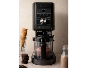 Ninja Creami machine in action, blending the chocolate mixture. A glass of ice cream cones and a bottle of chocolate milk are placed beside the machine.