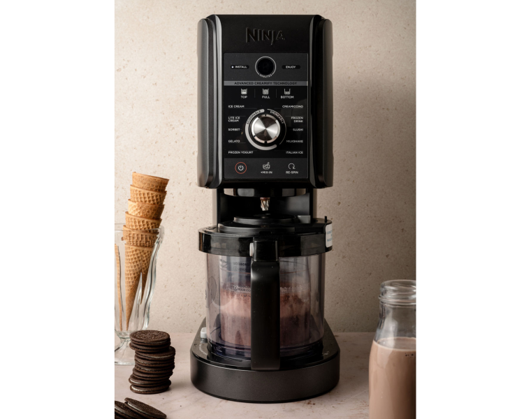 Ninja Creami machine in action, blending the chocolate mixture. A glass of ice cream cones and a bottle of chocolate milk are placed beside the machine.