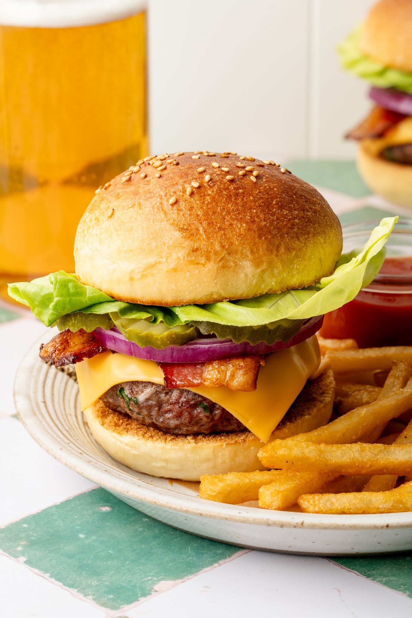 Close-up of a classic cheeseburger on a sesame seed bun, layered with lettuce, red onion, pickles, bacon, and cheddar cheese, served with golden fries and a pint of beer in the background.
