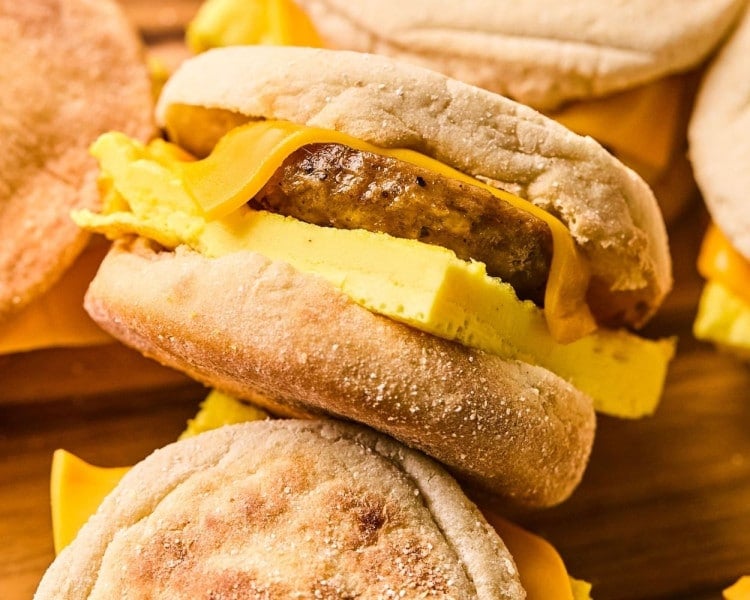 A close-up of a finished breakfast sandwich made with an English muffin, cheddar cheese, a sausage patty, and a thick square of baked egg. The sandwich is slightly tilted, showing the fluffy texture of the egg and the melted cheese.