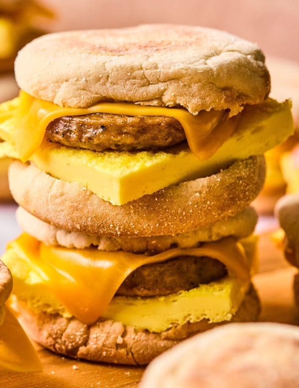 A close-up of a stacked pair of breakfast sandwiches made with English muffins, cheddar cheese, sausage patties, and thick folded egg. The cheese is slightly melted, and the sandwiches are set against a soft pink background.