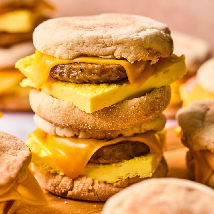 A close-up of a stacked pair of breakfast sandwiches made with English muffins, cheddar cheese, sausage patties, and thick folded egg. The cheese is slightly melted, and the sandwiches are set against a soft pink background.