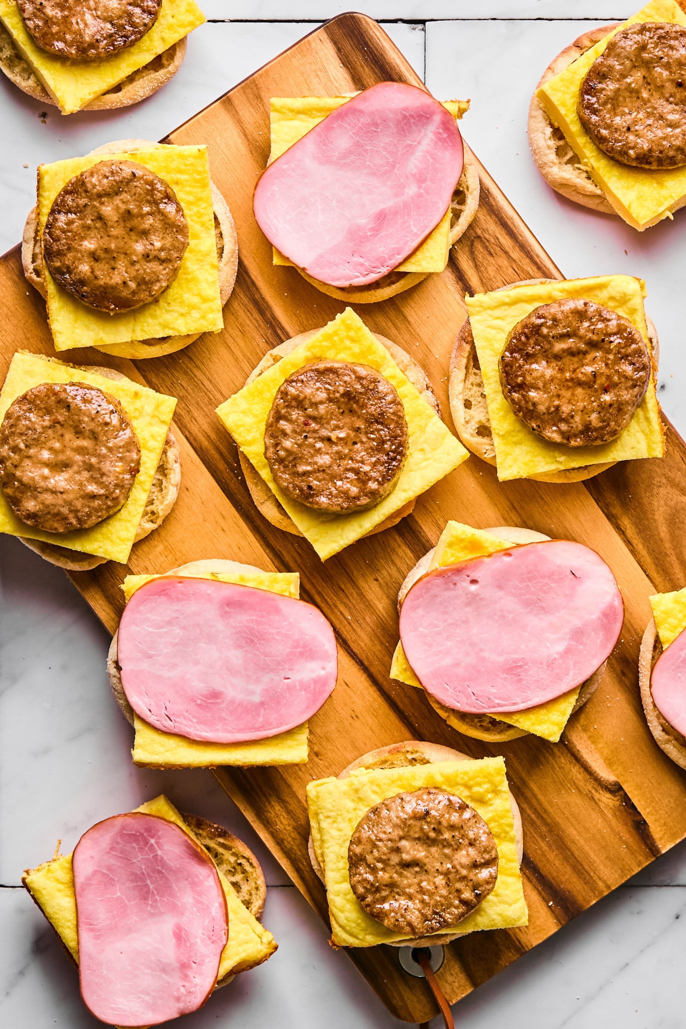 A wooden board topped with open-faced breakfast sandwiches in the process of being assembled. Each English muffin half is layered with a square of baked egg, and topped with either a round sausage patty or a slice of Canadian bacon. The arrangement shows a mix of the two meat options across the sandwiches.