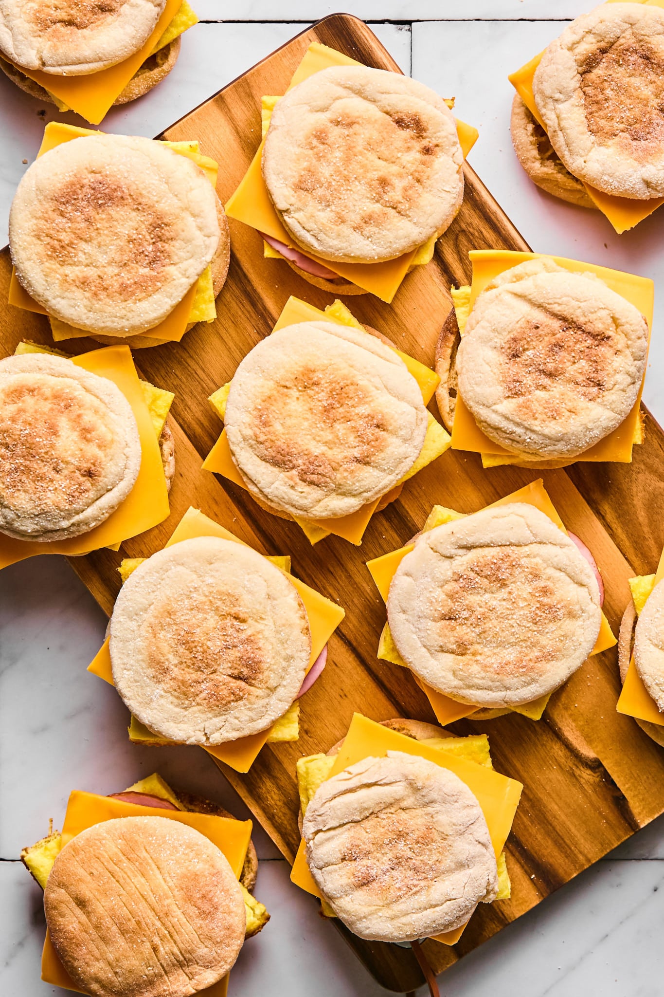 A wooden cutting board topped with multiple breakfast sandwiches made with English muffins, cheddar cheese, folded egg, and slices of ham. The sandwiches are arranged in a neat, overlapping pattern.