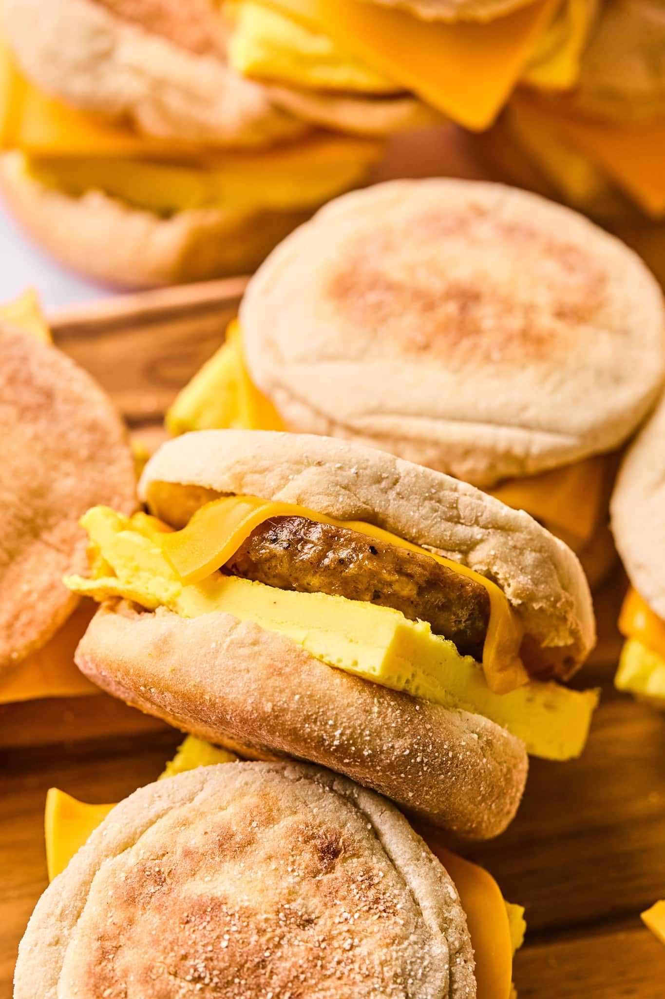 A close-up of a single breakfast sandwich made with an English muffin, cheddar cheese, a sausage patty, and folded egg. The sandwich is tilted, resting on top of other sandwiches on a wooden surface.