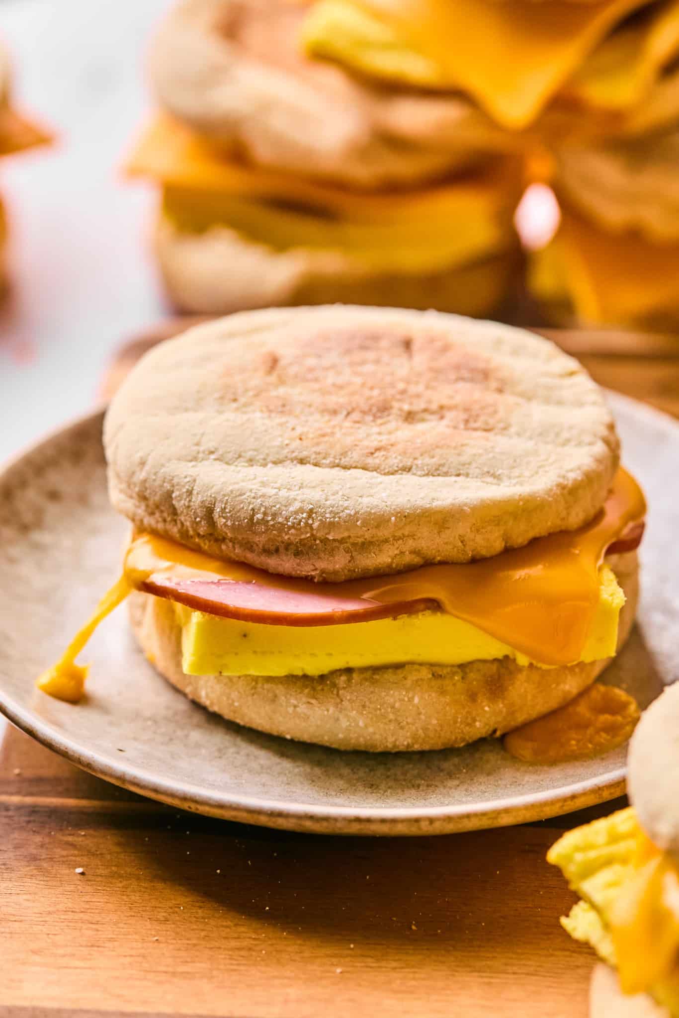 A breakfast sandwich on a ceramic plate made with an English muffin, cheddar cheese, folded egg, and a round slice of ham. The cheese is melted and slightly dripping from the edge.