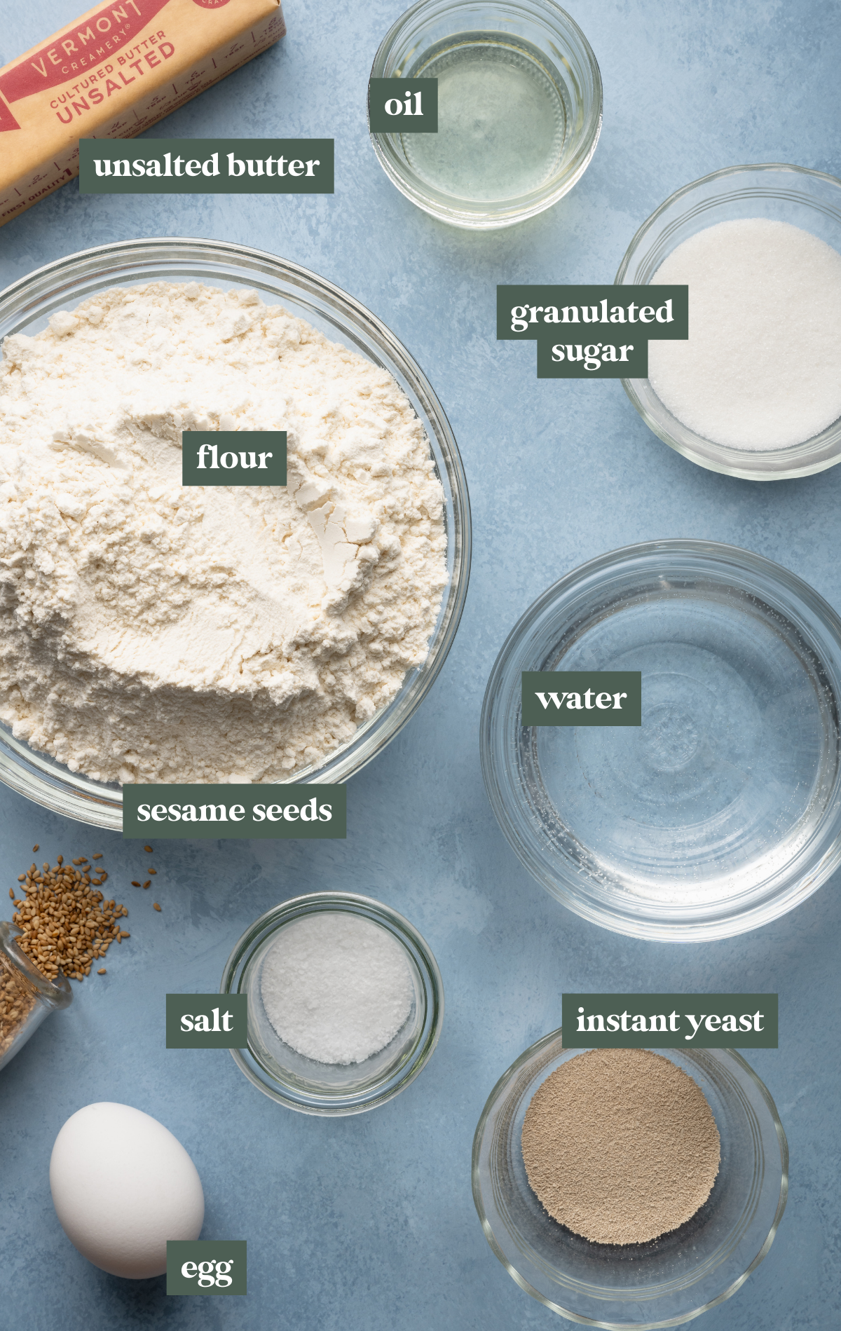 Flat lay of homemade hamburger bun ingredients including a bowl of flour, a stick of unsalted butter, granulated sugar, vegetable oil, yeast, salt, water, an egg, and a small jar of sesame seeds on a light blue surface.