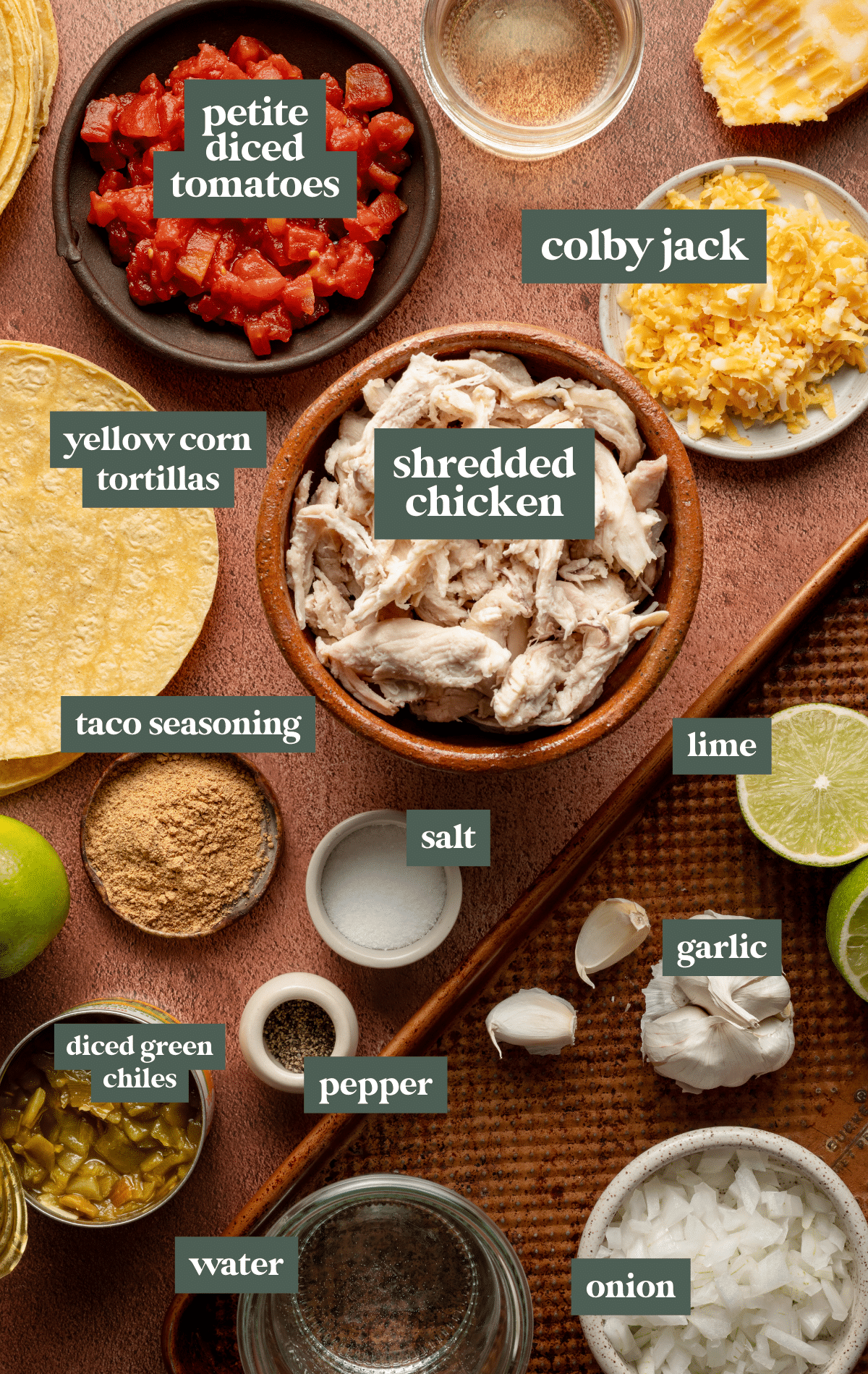 Flat lay of taco ingredients including shredded rotisserie chicken, taco seasoning, petite diced tomatoes, shredded Colby Jack cheese, canned green chiles, garlic, onion, lime, tortillas, and seasonings.
