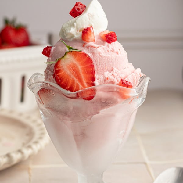Frozen strawberry yogurt in glass container topped with fresh strawbrries and whipped cream and spoon beside it.