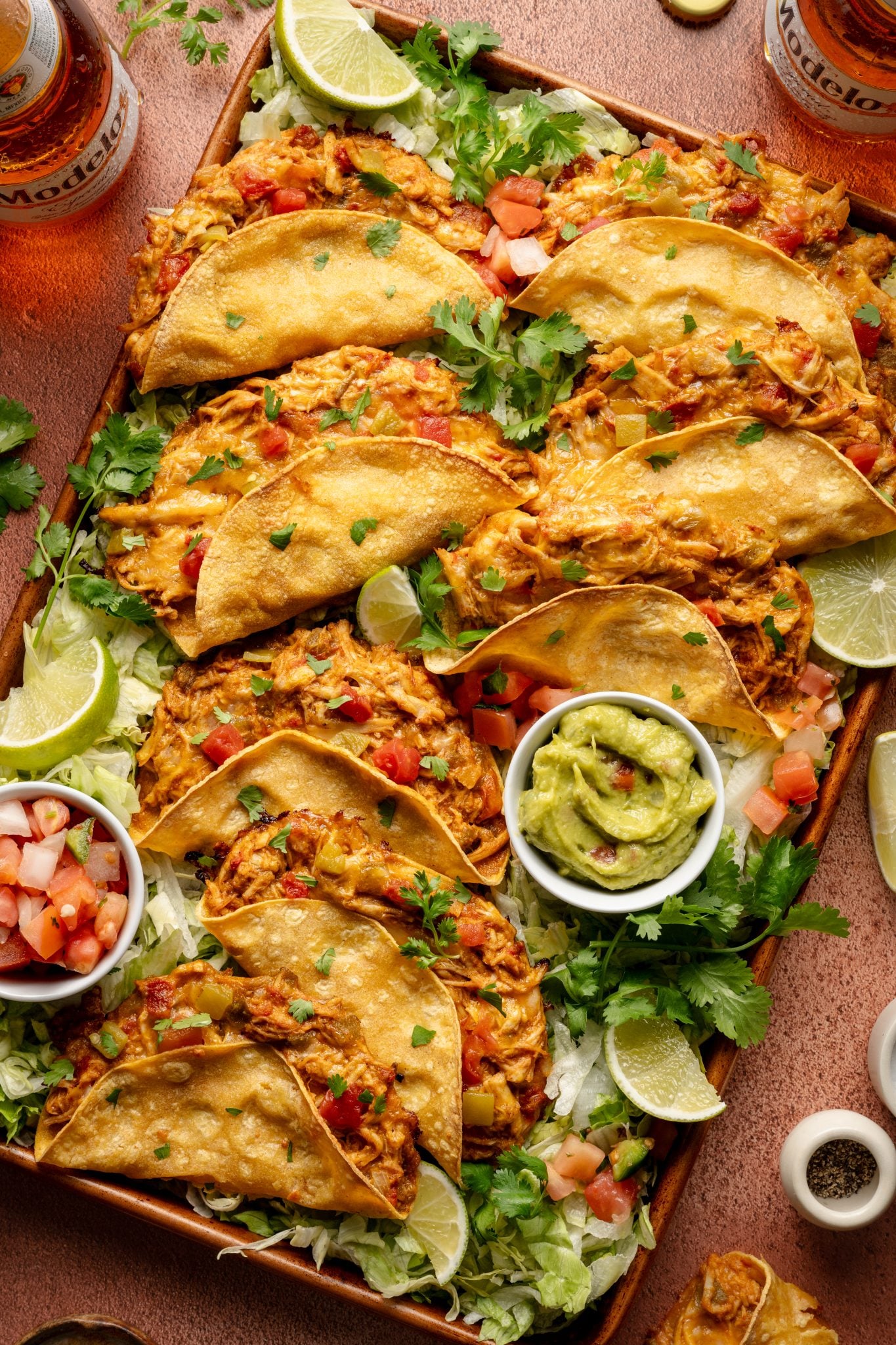 A sheet pan filled with golden baked chicken tacos, garnished with fresh cilantro and lime wedges. The tacos are served over a bed of shredded lettuce with sides of guacamole and pico de gallo.