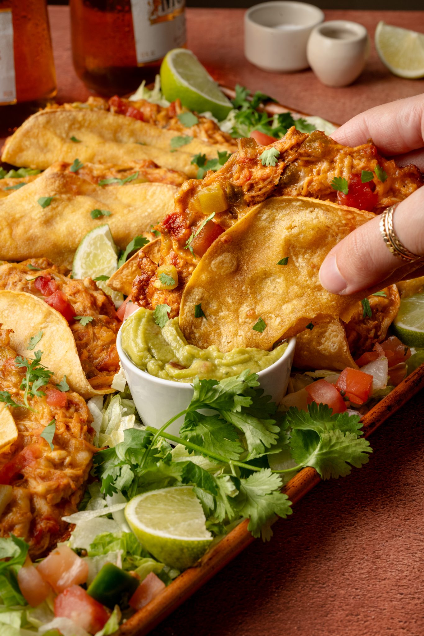 A hand picking up a baked chicken taco filled with cheesy shredded chicken, dipping it into a bowl of creamy guacamole. Tacos are arranged on a tray surrounded by cilantro, lettuce, and lime wedges.