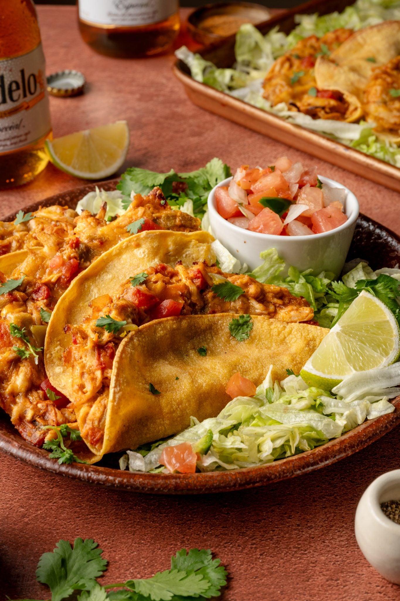 A close-up of a plate with three crispy baked chicken tacos filled with cheesy shredded chicken, resting on a bed of shredded lettuce and garnished with fresh cilantro. A lime wedge and a small bowl of fresh pico de gallo are served on the side, with bottles of Modelo beer and a tray of additional tacos in the background.