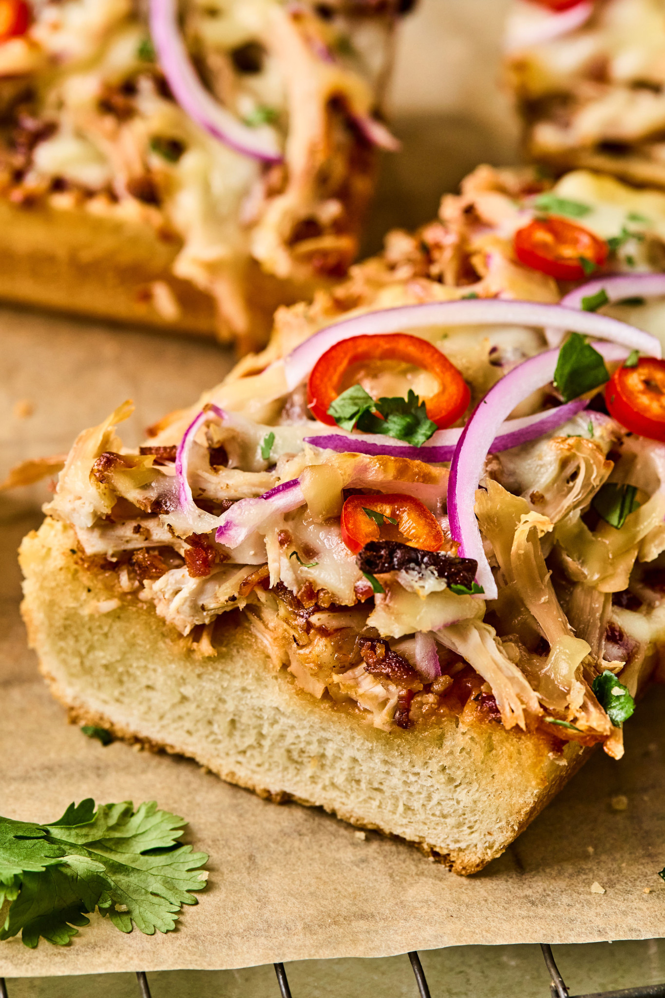Close-up of a slice of BBQ chicken French bread pizza topped with melted cheese, barbecue sauce drizzle, red onion, and sliced red chili peppers.

