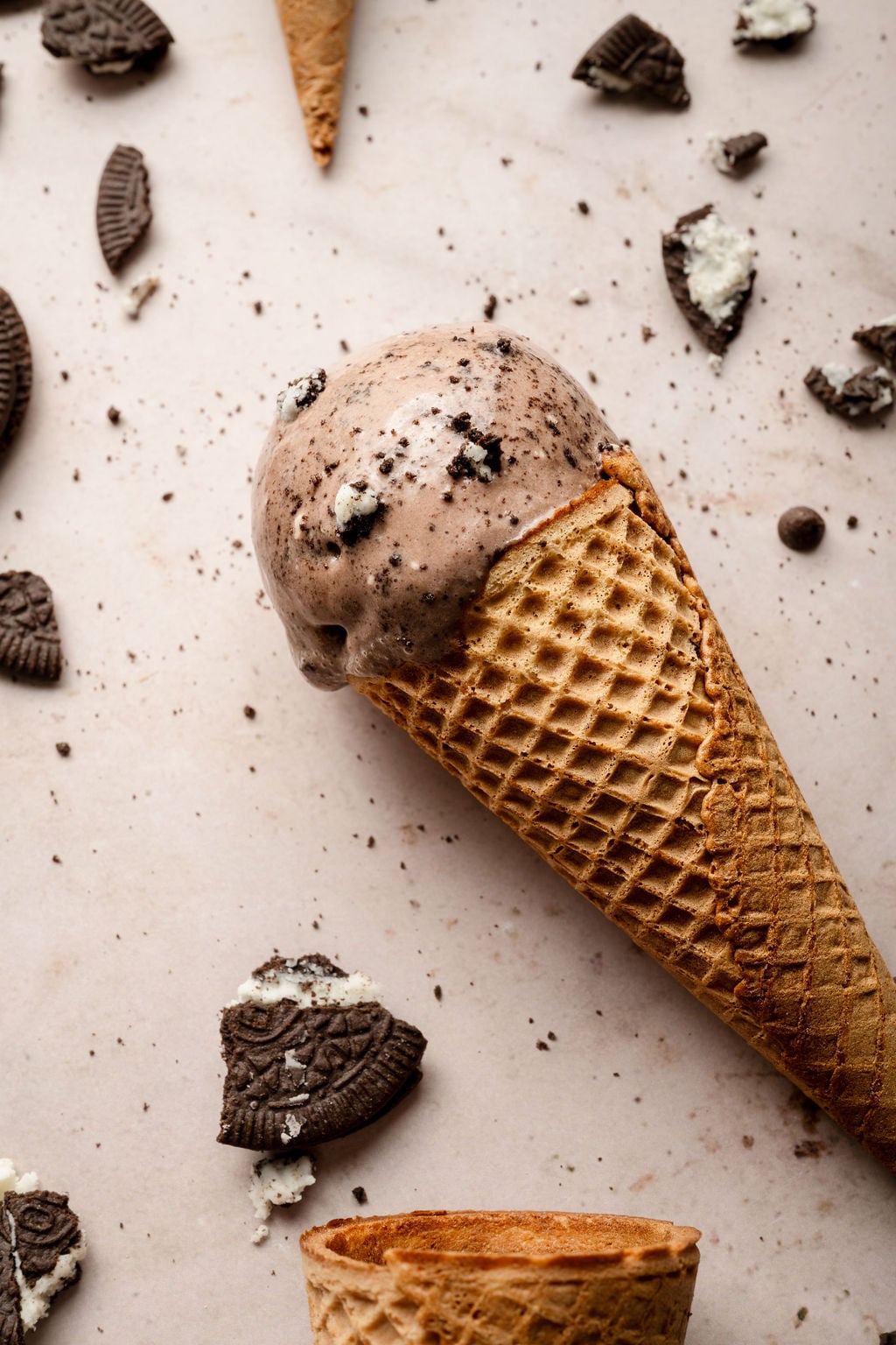 A scoop of chocolate Oreo ice cream in a waffle cone, lying on a light surface scattered with crumbled Oreo cookie pieces and chocolate chips.