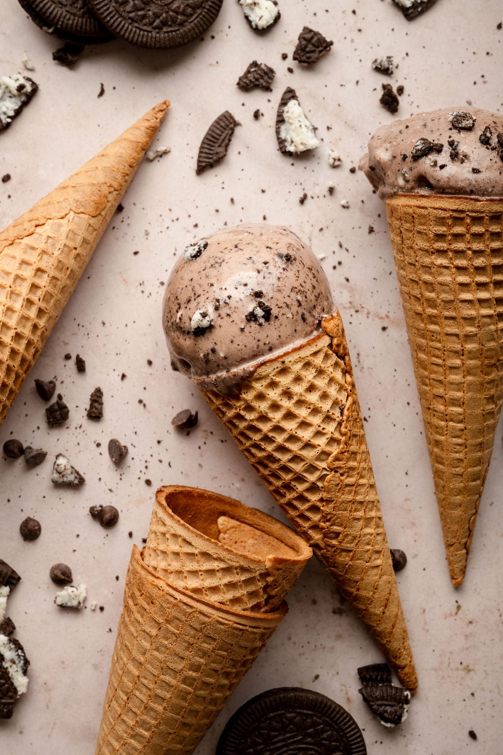 Overhead view of two waffle cones filled with scoops of chocolate Oreo ice cream, surrounded by additional empty cones, chocolate chips, and crumbled Oreo pieces on a light surface. The ice cream is speckled with bits of crushed cookies.