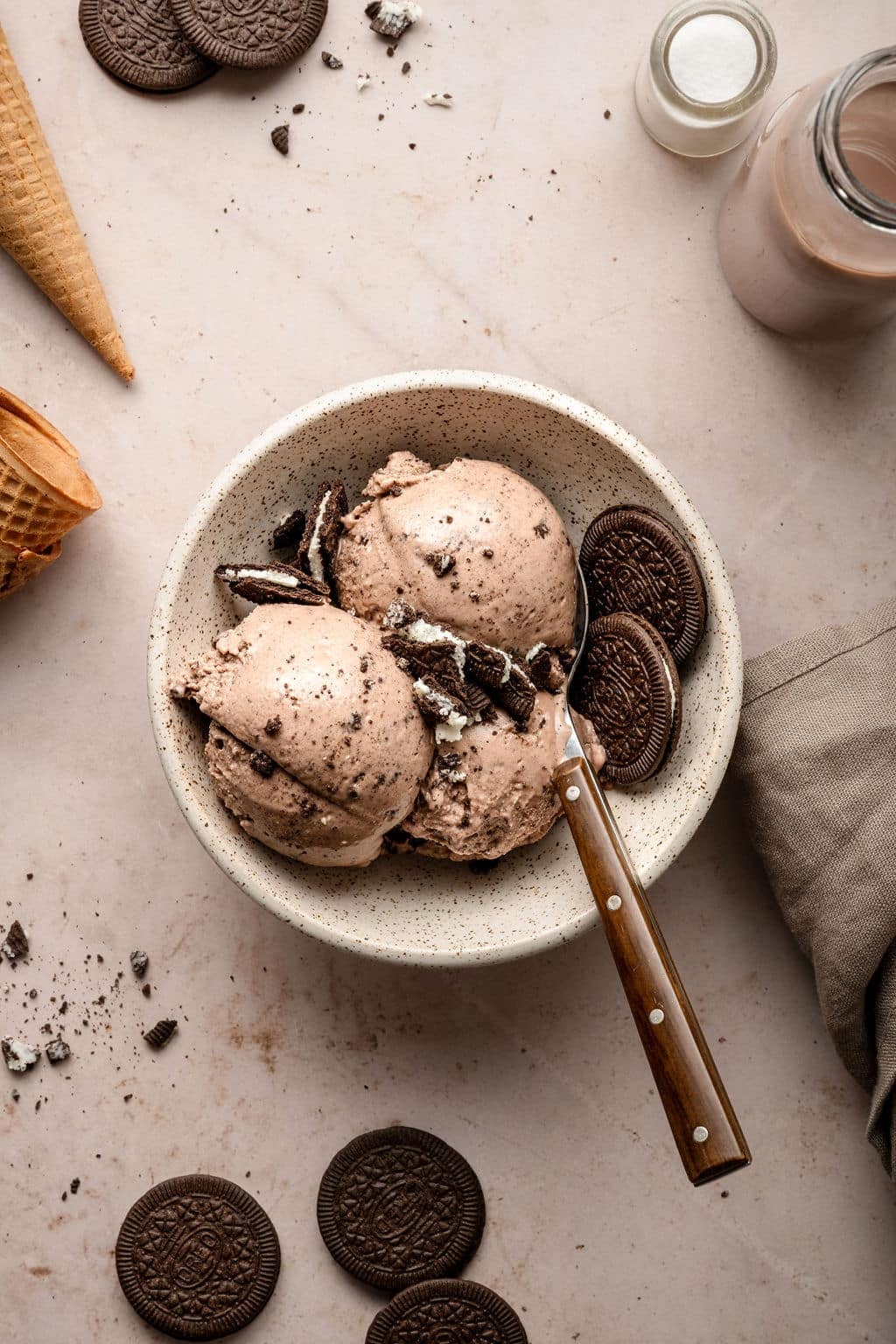 Overhead view of a speckled bowl filled with three scoops of chocolate Oreo ice cream, garnished with crushed and whole Oreos. The scene is surrounded by cones, cookies, and a jar of chocolate milk.