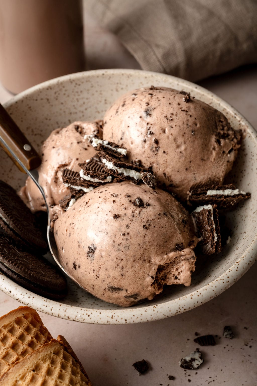 Angled view of a bowl of chocolate Oreo protein ice cream with large scoops and cookie crumbles on top, placed beside waffle cones and a glass of chocolate milk.