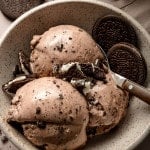 A speckled ceramic bowl filled with three scoops of creamy chocolate Oreo ice cream, topped with crushed Oreo cookie pieces. Whole Oreo cookies are placed on the side and in the background, with a metal spoon resting in the bowl. The ice cream is smooth with visible cookie bits throughout.