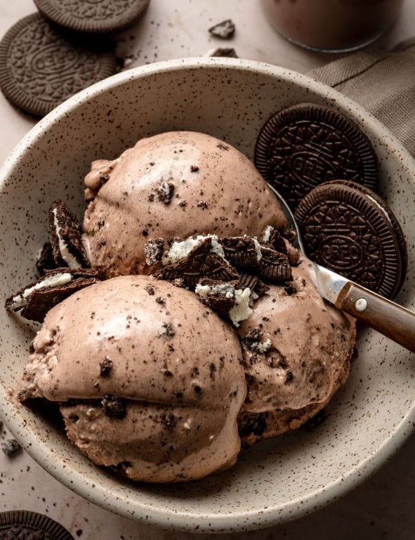 A speckled ceramic bowl filled with three scoops of creamy chocolate Oreo ice cream, topped with crushed Oreo cookie pieces. Whole Oreo cookies are placed on the side and in the background, with a metal spoon resting in the bowl. The ice cream is smooth with visible cookie bits throughout.
