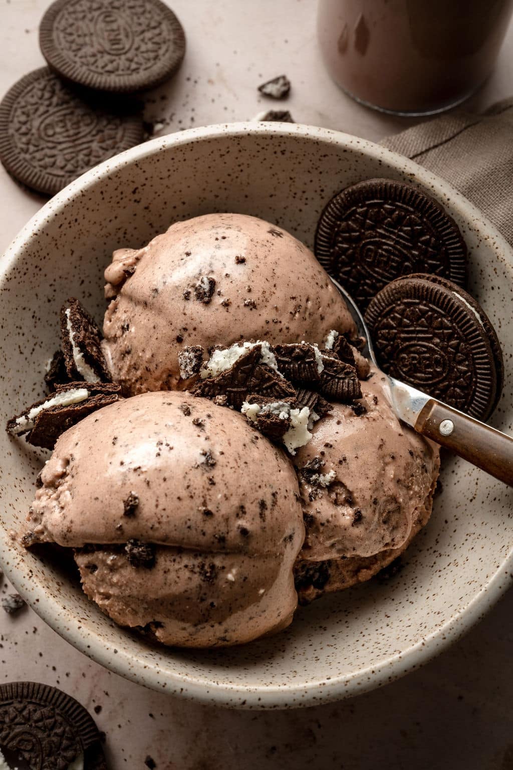 A speckled ceramic bowl filled with three scoops of creamy chocolate Oreo ice cream, topped with crushed Oreo cookie pieces. Whole Oreo cookies are placed on the side and in the background, with a metal spoon resting in the bowl. The ice cream is smooth with visible cookie bits throughout.