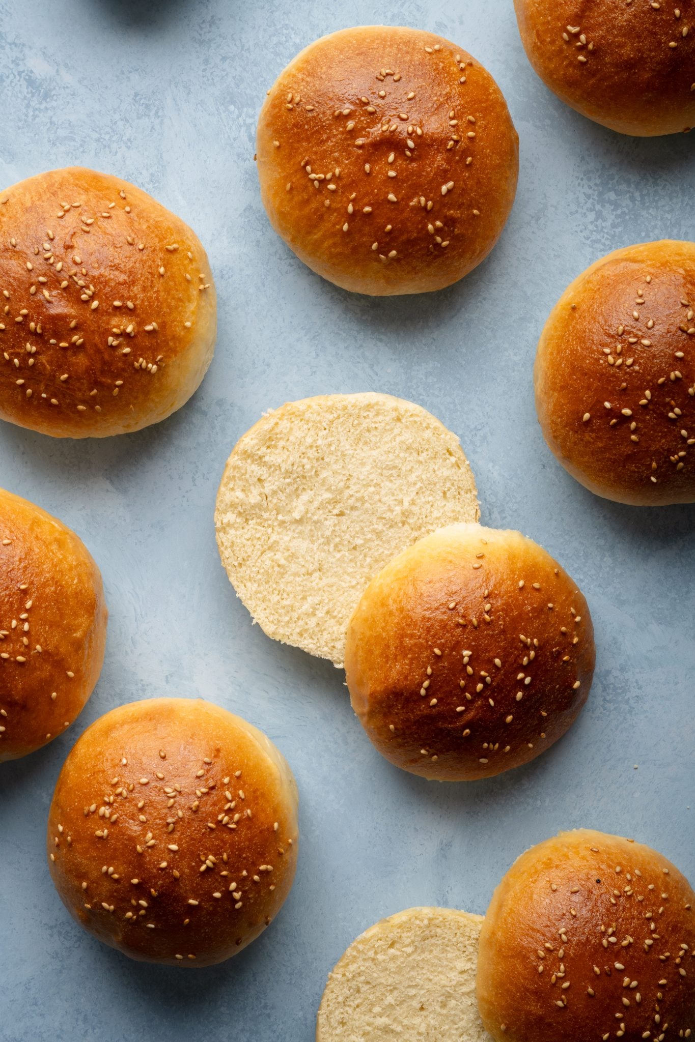 Top-down view of several golden hamburger buns with one sliced open to reveal its soft, fluffy interior.