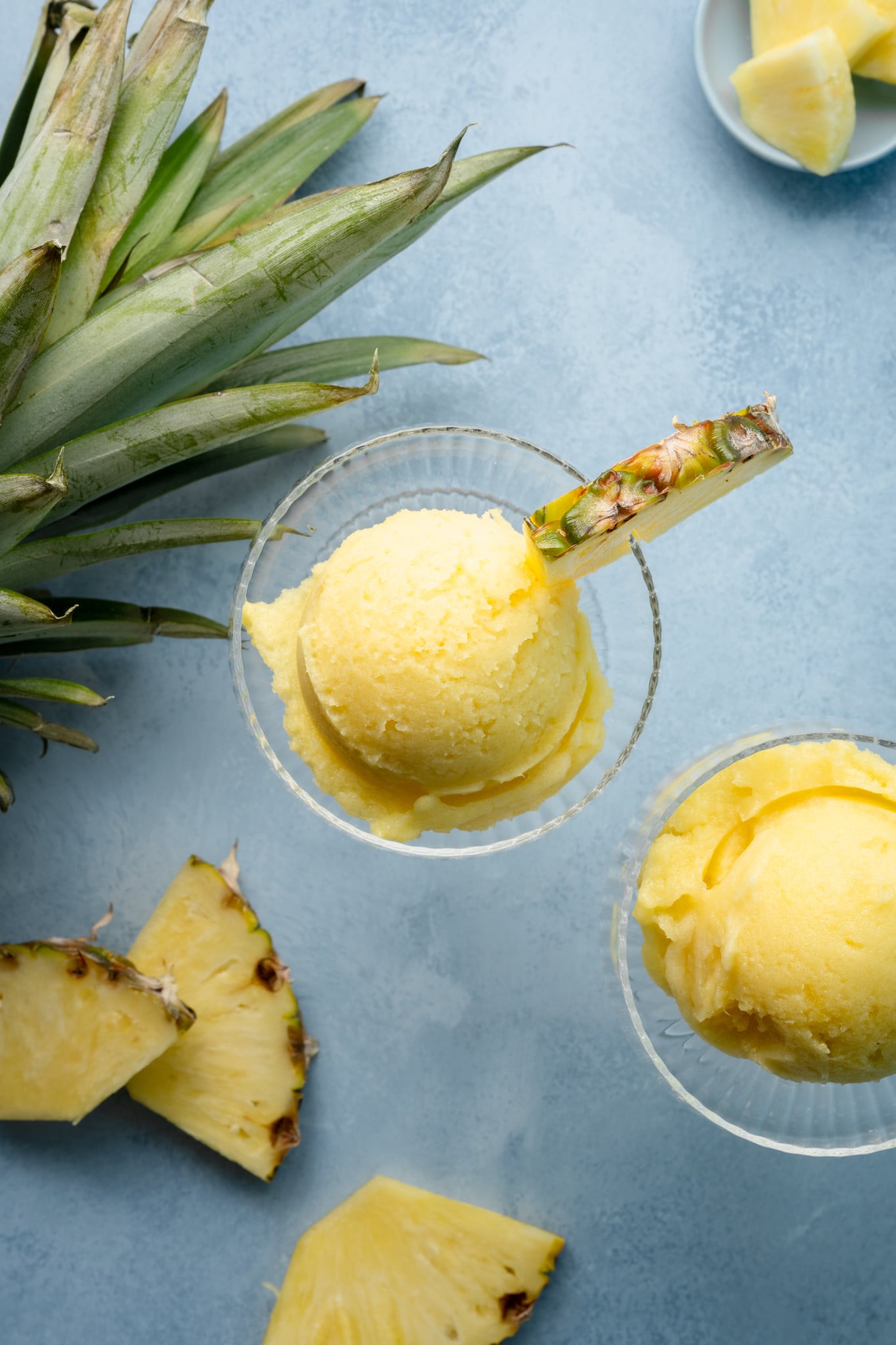 Overhead view of two scoops of creamy pineapple sorbet in glass dessert cups, each garnished with a fresh pineapple wedge, surrounded by pineapple leaves and slices.

