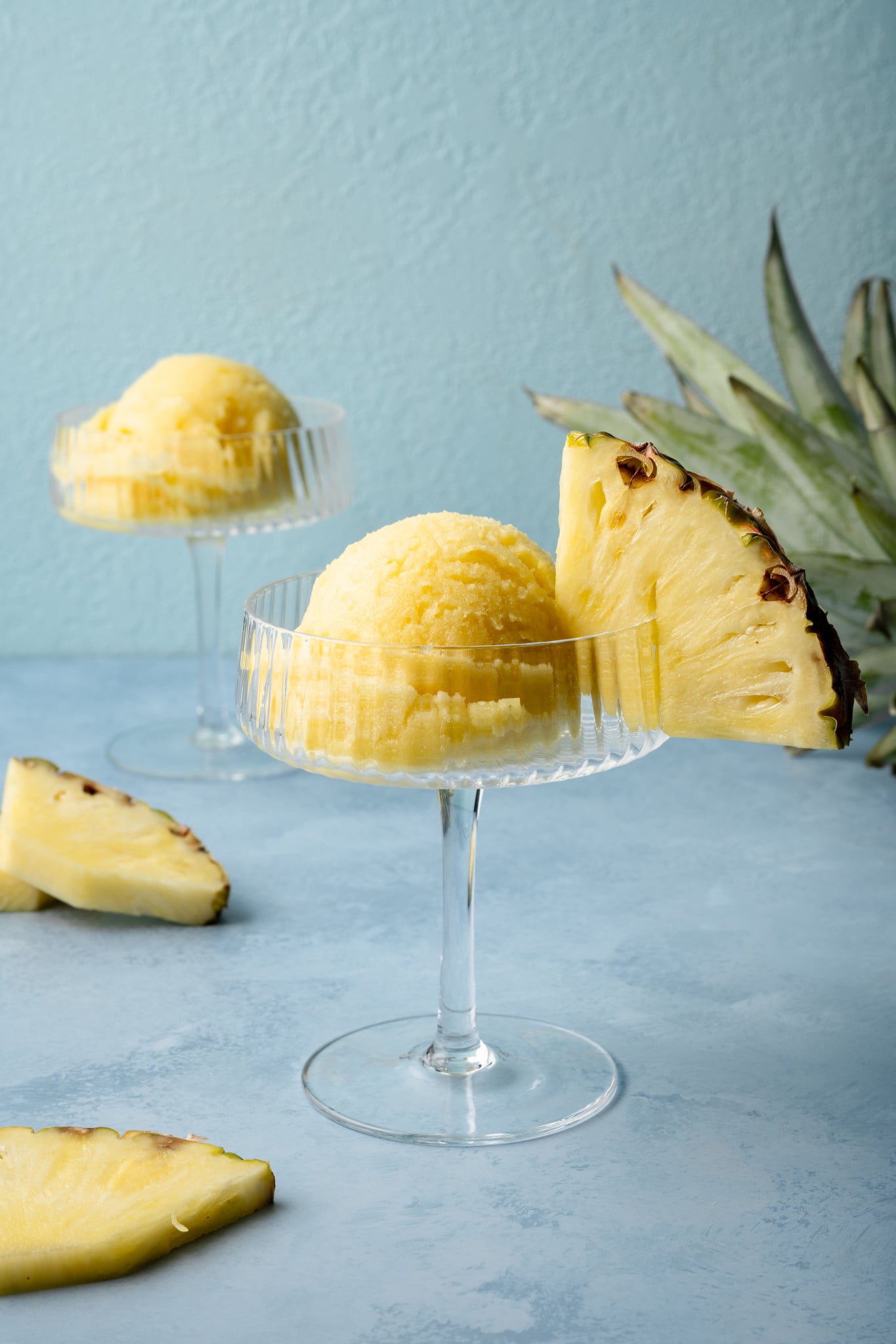 Close-up of pineapple sorbet served in a clear coupe glass with a thick pineapple wedge on the rim, placed on a blue background with a pineapple crown nearby.

