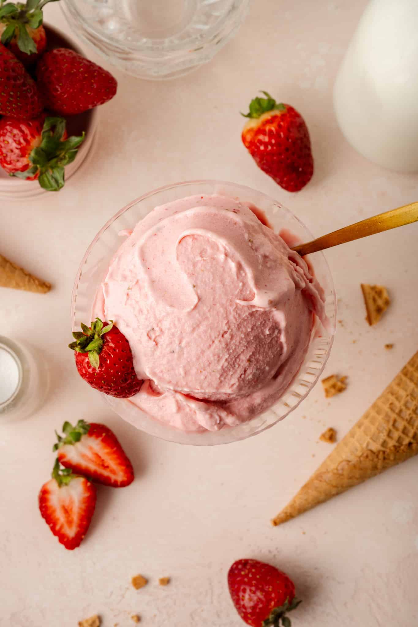 Overhead shot of strawberry ice cream in glass bowl with strawberries and sugar cone on the table below glass and gold spoon.