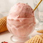 Strawberry ice cream in short glass bowl with gold spoon and waffle cones surrounding it on the table.