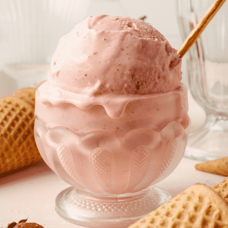 Strawberry ice cream in short glass bowl with gold spoon and waffle cones surrounding it on the table.