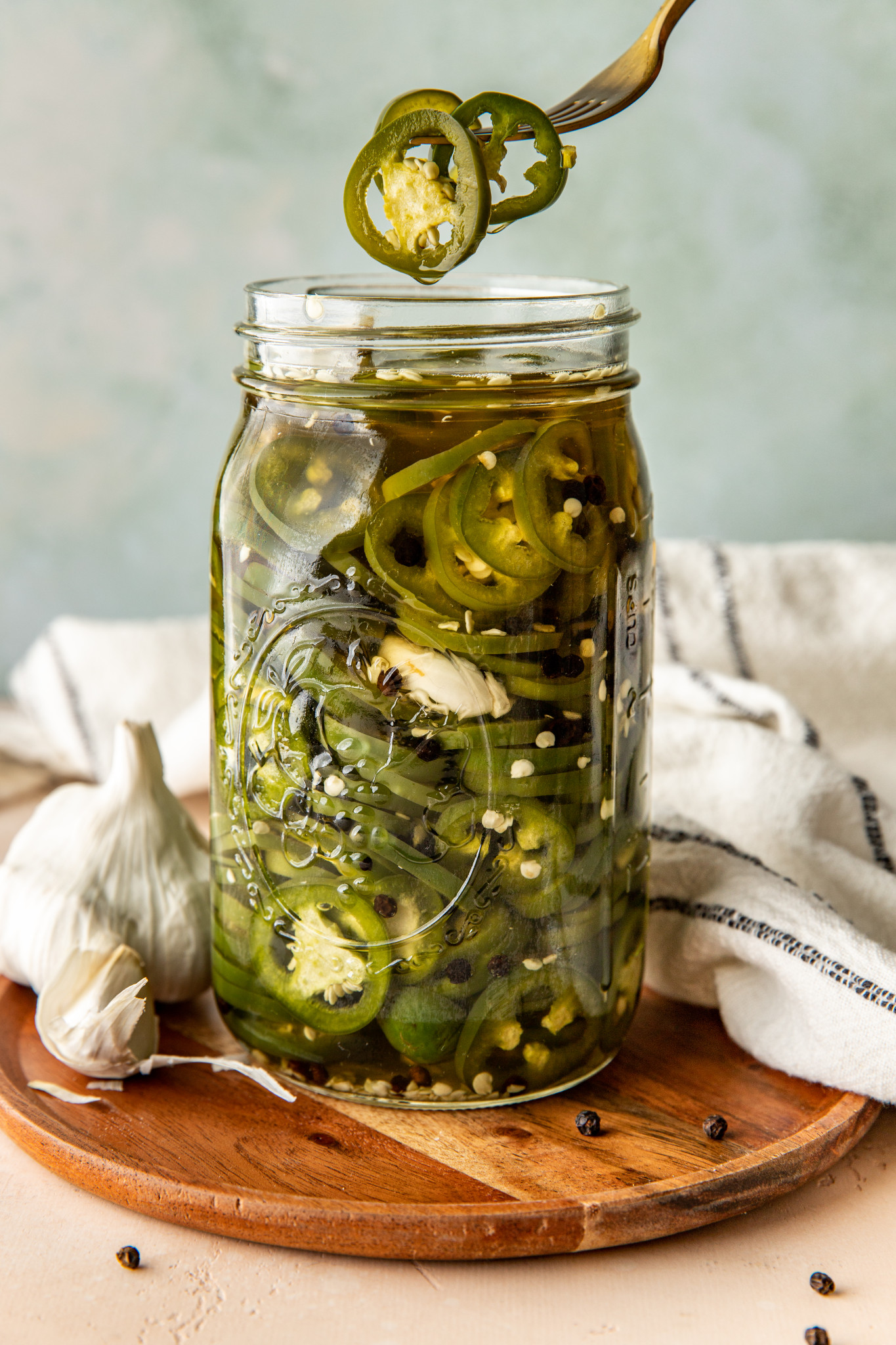 a fork picking up 3 pickled jalapeños from a glass mason jar sitting on a circular wooden platter.