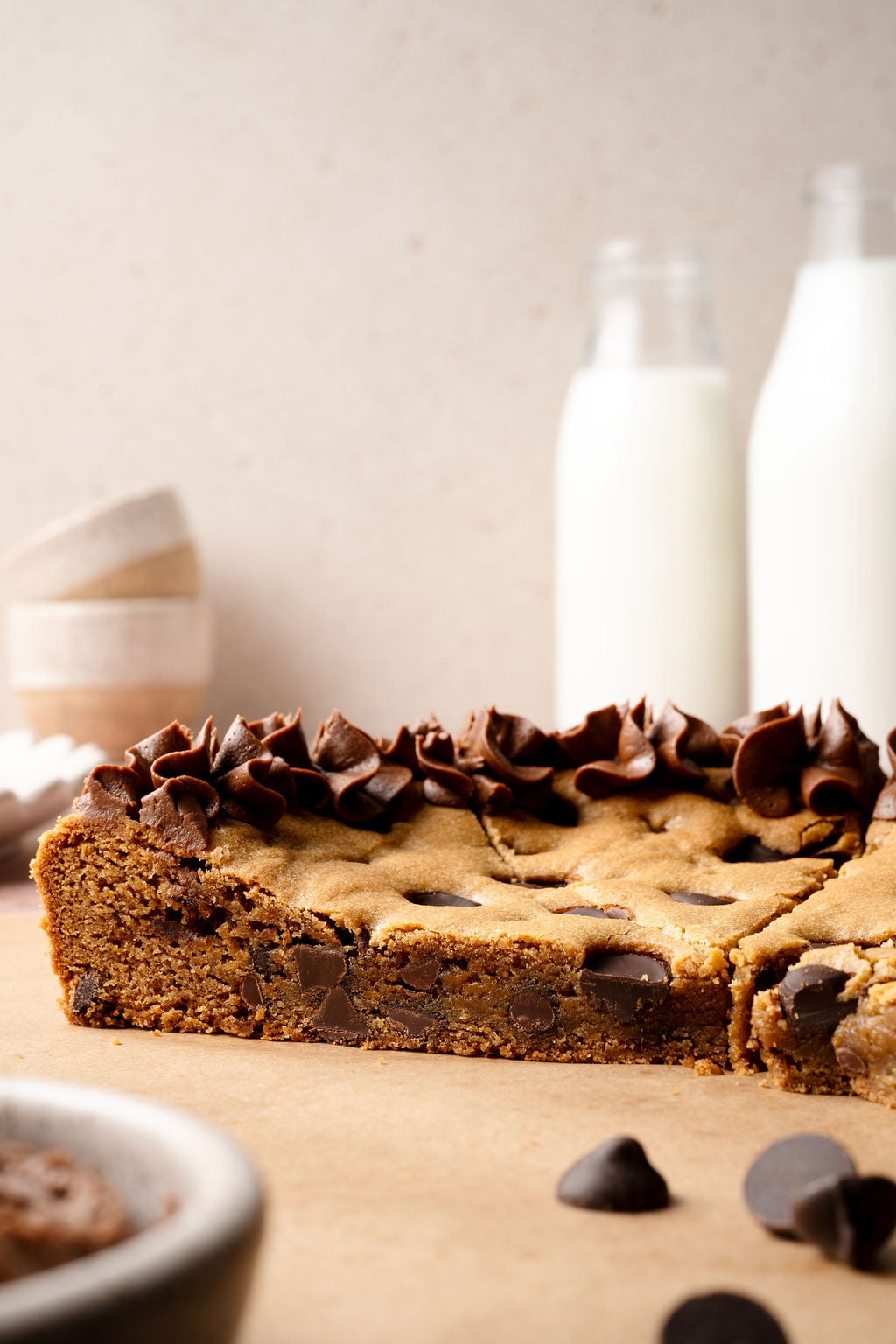 A side view of the chocolate chip cookie cake showing the thick, chewy interior filled with chocolate chips, topped with decorative chocolate frosting and two bottles of milk in the background.