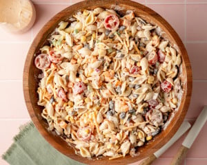 Overhead view of fully mixed creamy taco pasta salad in a large wooden bowl. Shell pasta, cherry tomatoes, corn, black beans, shredded cheese, and diced veggies are coated in the creamy taco dressing.