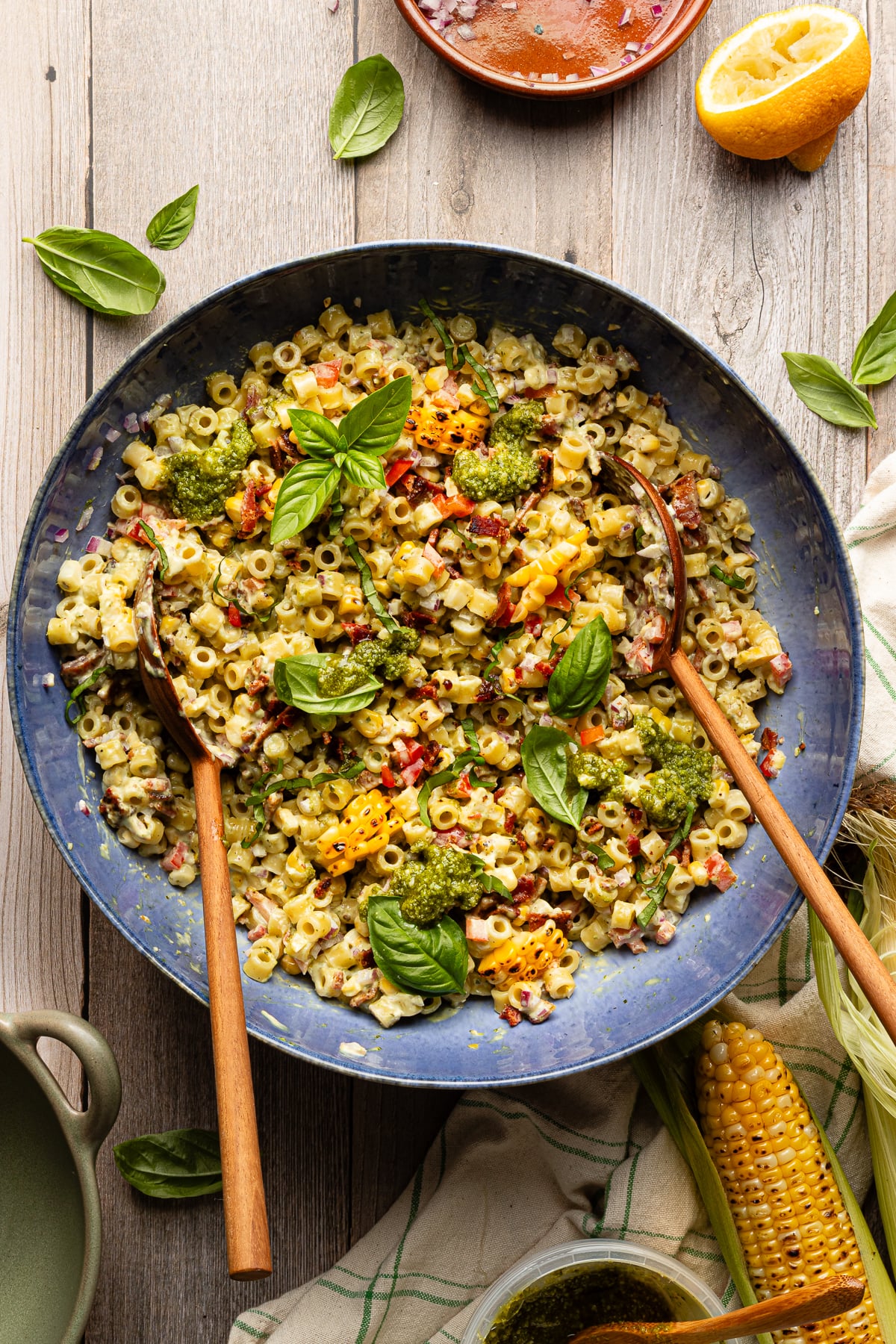 Pesto pasta salad tossed together in large blue serving bowl with wood spoons and garnished with fresh basil.